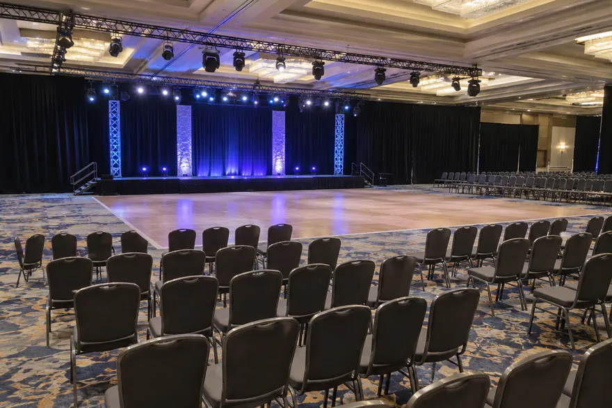 A hotel ballroom set up for a show: a lit stage with a wooden dance floor in front and rows of empty chairs.
